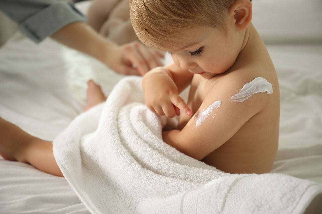 Bebé envuelto en una toalla blanca señalando crema humectante aplicada en su brazo.